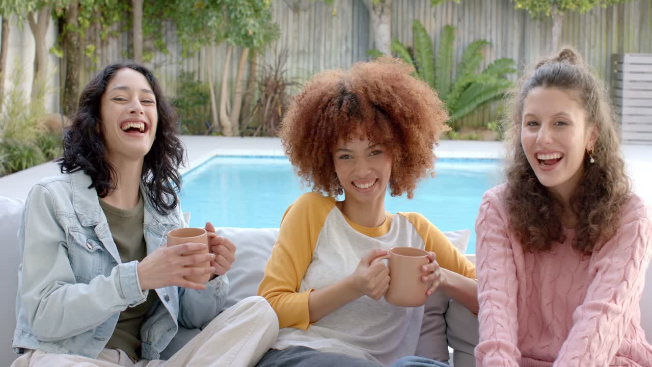 Diverse female friends sharing coffee on sofa by pool and laughing after punchline lifting mugs