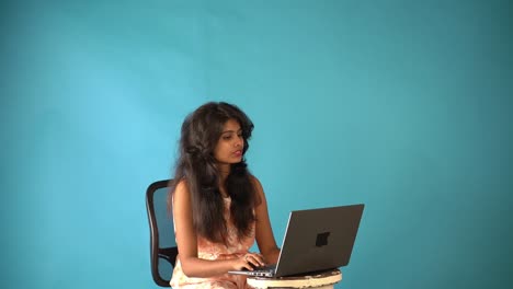 Ein-Junges-Indisches-Mädchen-In-Orangefarbenem-Kleid,-Das-Einen-Videoanruf-Mit-Einem-Laptop-Macht,-Der-Auf-Einem-Isolierten-Blauen-Hintergrund-Sitzt