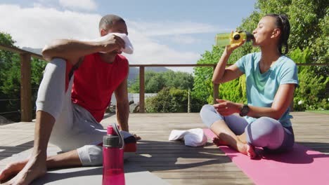 Tired-biracial-couple-on-terrace-drinking-water,-resting-after-exercises-and-talking