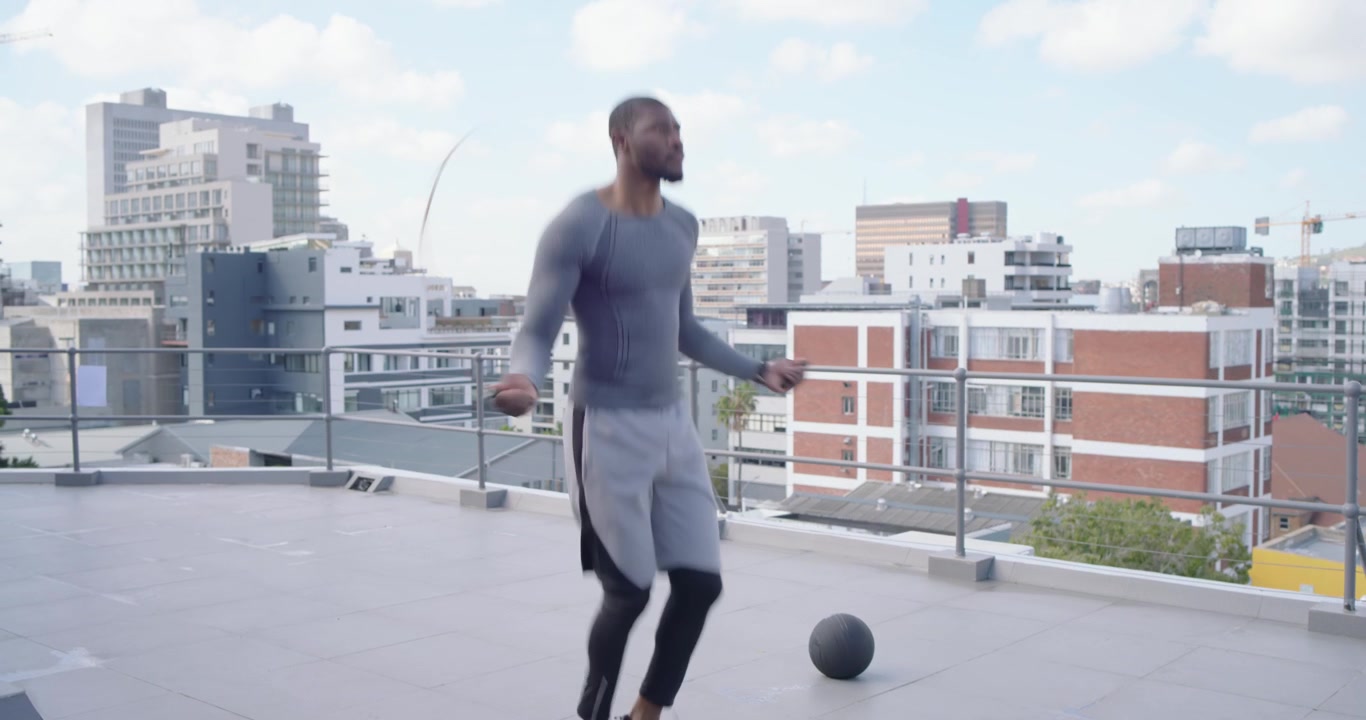 Pria Afrika memulai latihan di tengah, melompat tali di atap dengan pakaian abu-abu, bola medis di sebelah kanan untuk latihan kardio.