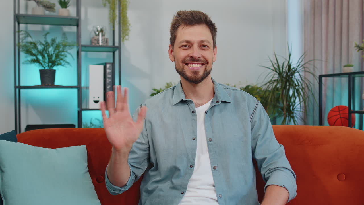 joven sonriendo amistosamente a la cámara, agitando las manos gestando hola, hola, saludando en casa en el sofá