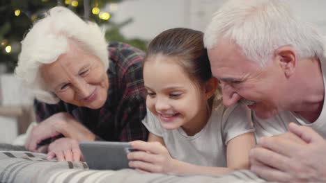 nonni felici con la loro nipotina sdraiata sul letto e che fa una videochiamata a natale