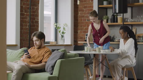 dos chicas desayunando juntas en casa mientras su compañero de habitación se sienta en el sofá y usa el teléfono