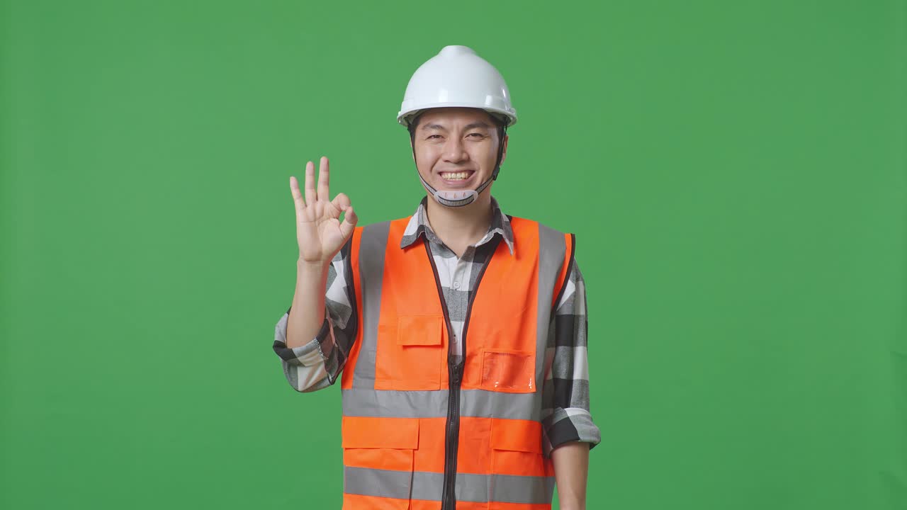 Asian Male Engineer With Safety Helmet Smiling And Showing Okay Gesture To The Camera While Standing In The Green Screen Background Studio