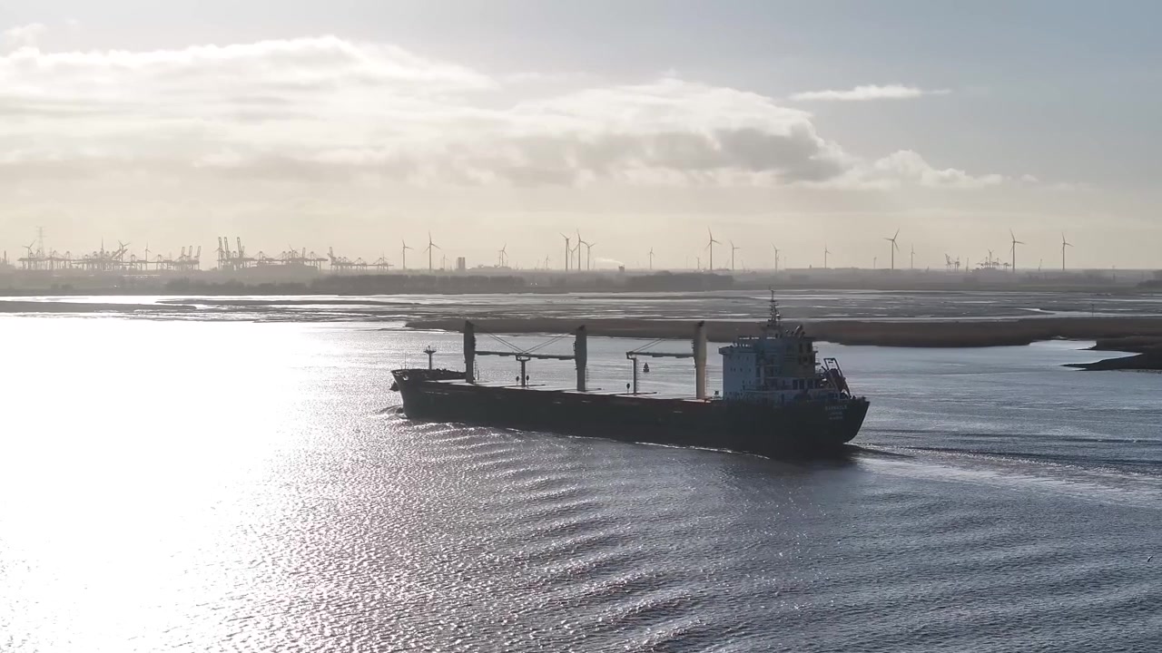 Imágenes de dron de un buque de carga navegando por el Escalda, con los humedales de Saeftinghe, grúas portuarias y aerogeneradores distantes visibles en un día soleado.