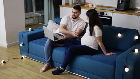 Hübsches-Schwangeres-Paar,-Das-Einen-Laptop-Benutzt-Und-Auf-Dem-Sofa-In-Einem-Neuen-Wohnzimmer-Mit-Girlanden-Sitzt