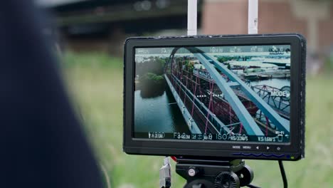 Handheld-camera-view-of-monitor-attached-to-tripod-showing-red,-arched-bridge-over-a-river,-trees-and-buildings-in-background,-with-settings-visible-on-display