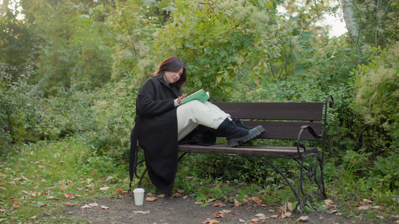Outdoor woman seated on bench with legs up, reading book in lush park, coffee paper cup on ground, calm autumn greenery around, cozy black coat and white trousers, quiet study moment
