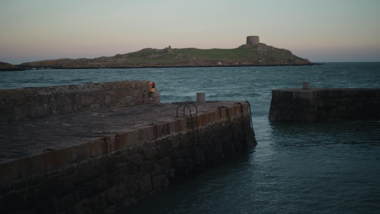 A locked-off shot of Colliemore Harbour pier in Dublin, Ireland, with Dalkey Island and Martello Tower in the background. Gentle waves move under a pastel sunset sky.