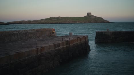 a locked-off shot of colliemore harbour pier in dublin, ireland, with dalkey island and martello tower in the background. gentle waves move under a pastel sunset sky.