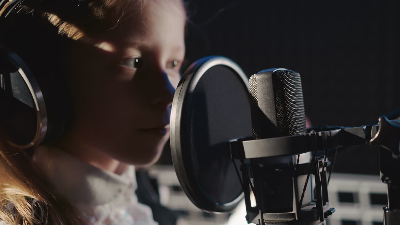 Kid touches microphone in recording studio. Inquisitive young girl in headphones inspects microphone by finger in acoustically isolated room