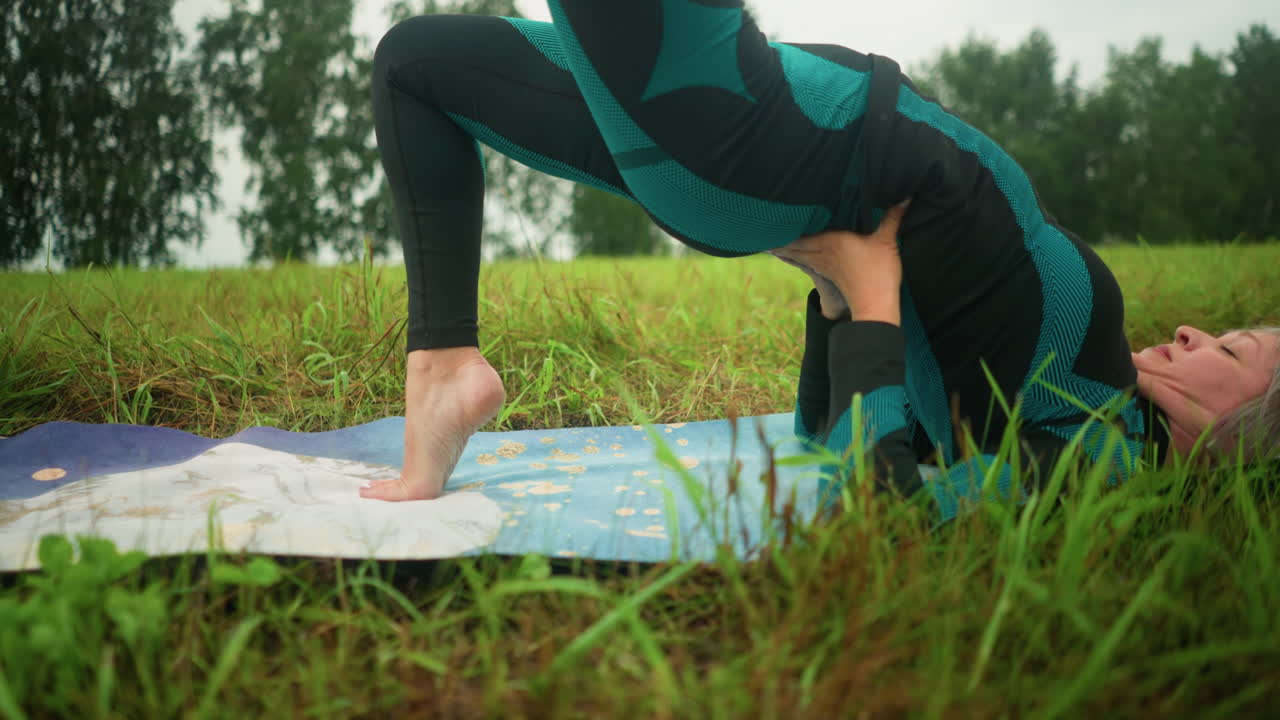 mujer de cabello gris acostada en una alfombra de yoga practicando la postura del puente con las manos apoyando su cintura, sentada en un vasto campo de hierba bajo un cielo nublado, con árboles en la distancia