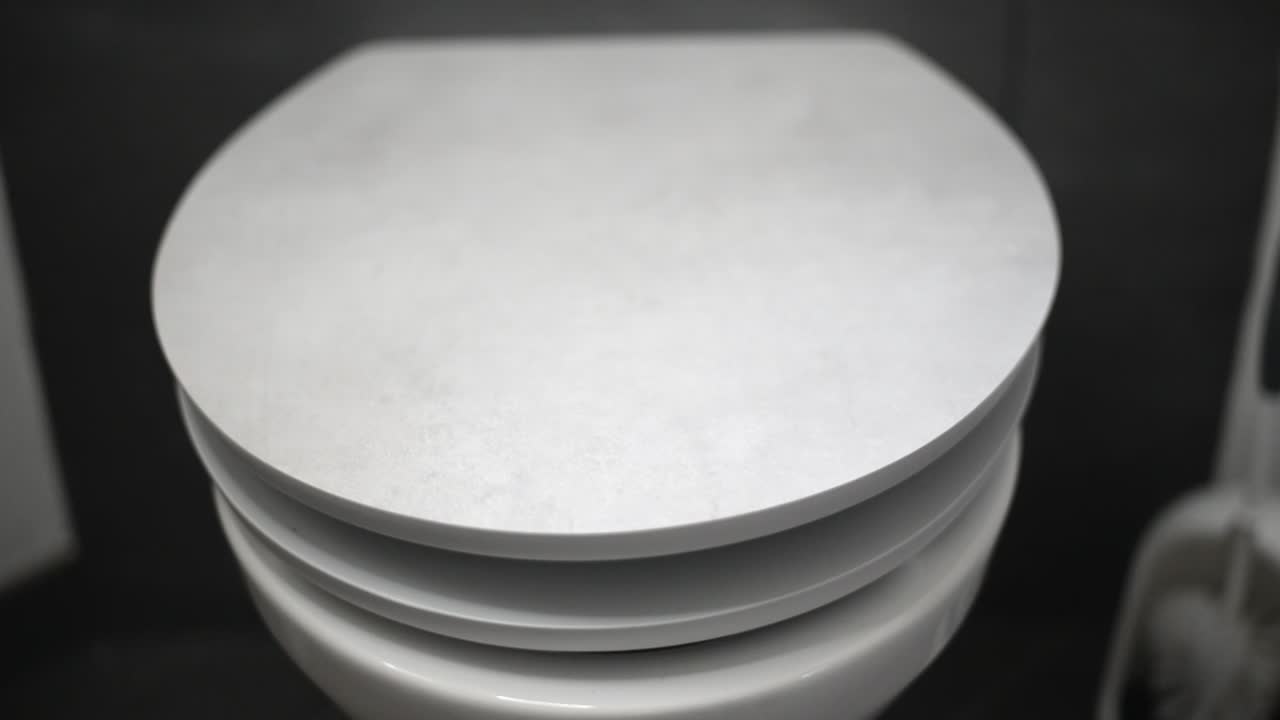 Soft close toilet seat closing in front of grey wall and white toilet brush in background