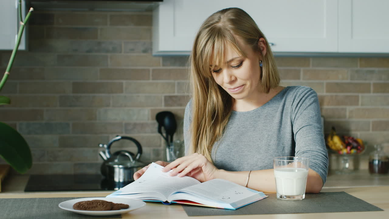 joven mujer atractiva leyendo un libro en la cocina cercana se encuentra un concepto de vaso de leche - saludable