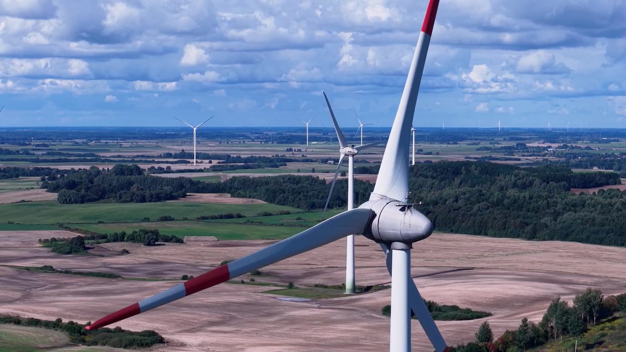 Flyfoto viser vindturbiner som roterer over et vidstrakt lappeteppe av jordbruksland, frodige åkre og skoger under en dynamisk skyfylt himmel, noe som fremhever ren energi i et levende landlig landskap