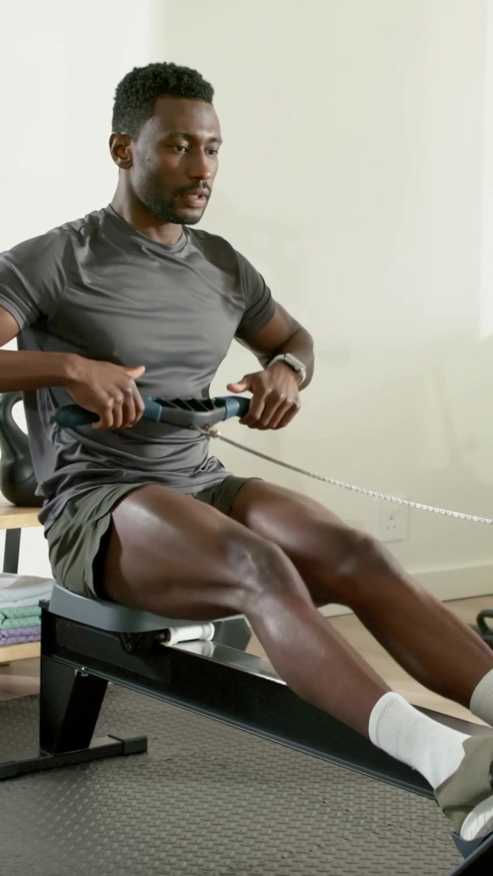 Vertical video: Rowing man on rower in grayT driving legs from catch, pulling for workout at home