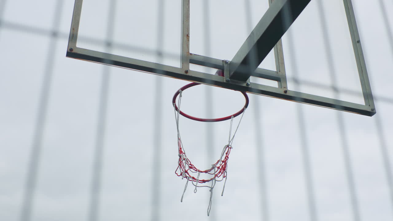 Canestro da basket con rete a catena su cielo nuvoloso, canestro arrugginito e tabellone in vetro incorniciato da una recinzione verticale, gocce di pioggia attaccate alle maglie, oscillazione al rallentatore, atmosfera da parco giochi vuoto con dettagli urbani