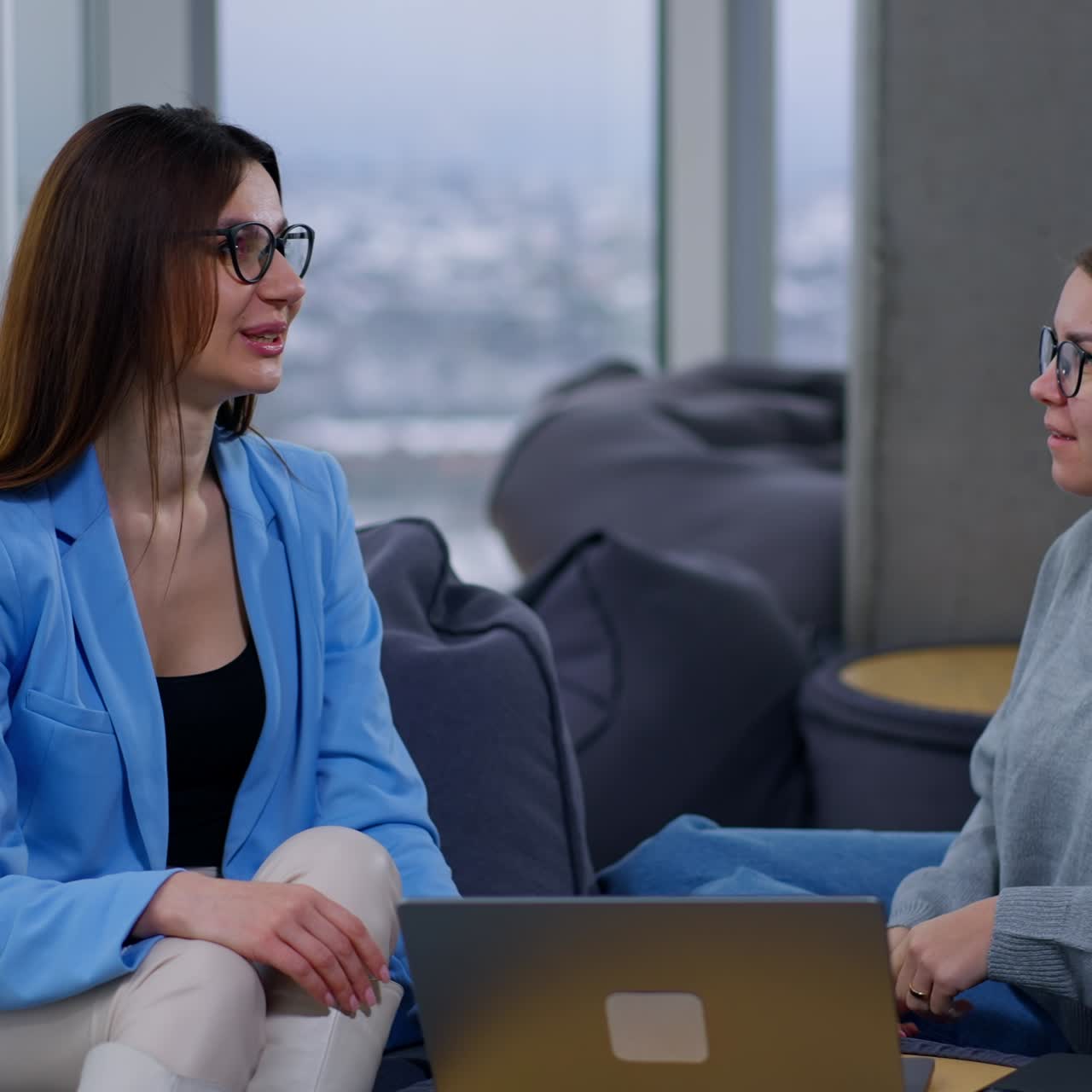Colleghe che lavorano in co-working sedute sul divano e discutono delle loro idee. Guardano il portatile, parlano e ridono comunicando