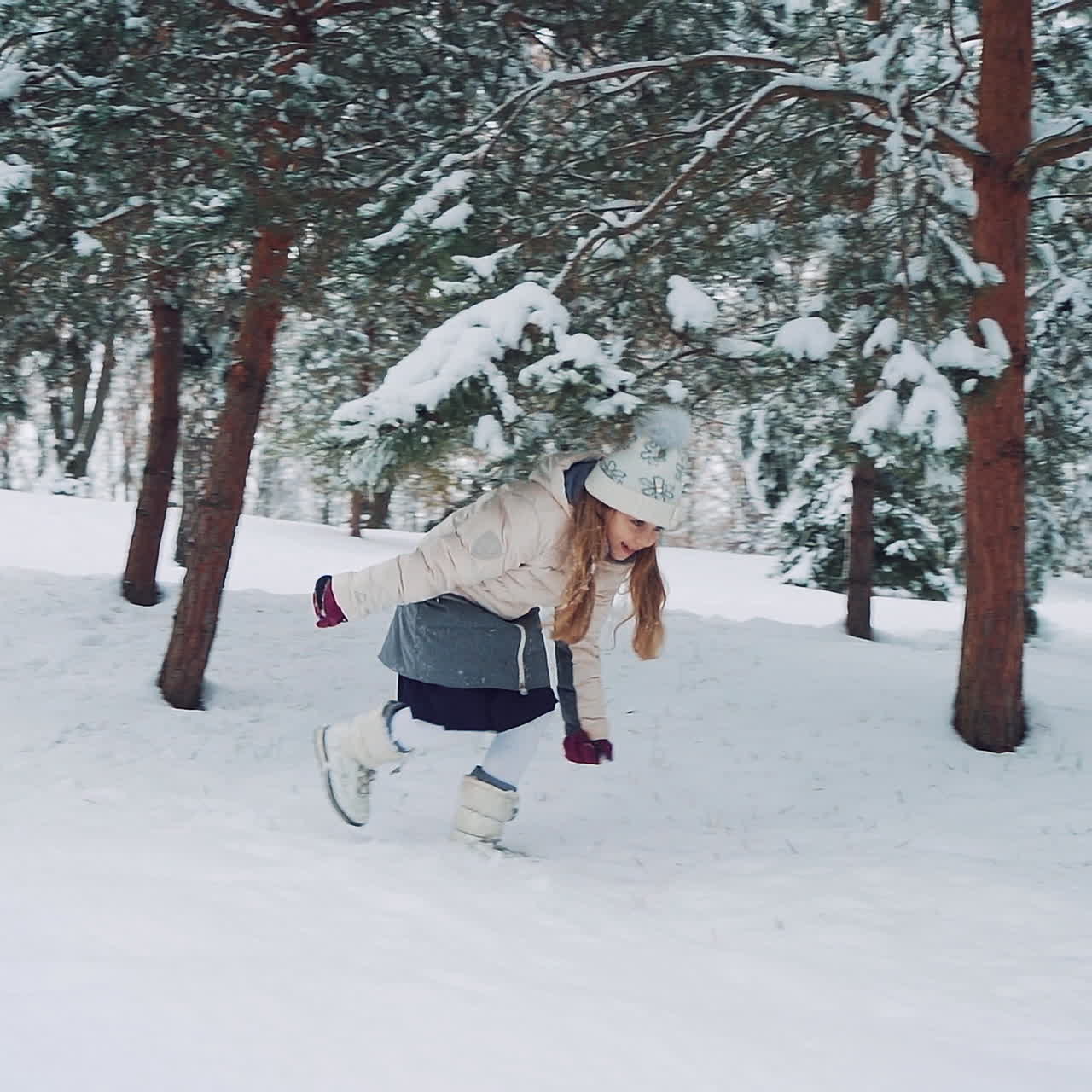 Cute little girl running in the snow among beautiful fir-trees in winter. One girl going to help her friend to raise up from white snow outdoors. Slow motion.
