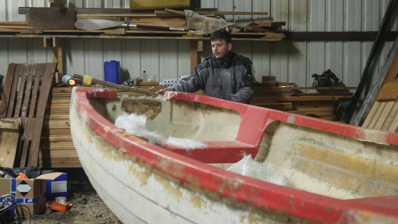 Man using heat gun on fiberglass boat repair in cold workshop