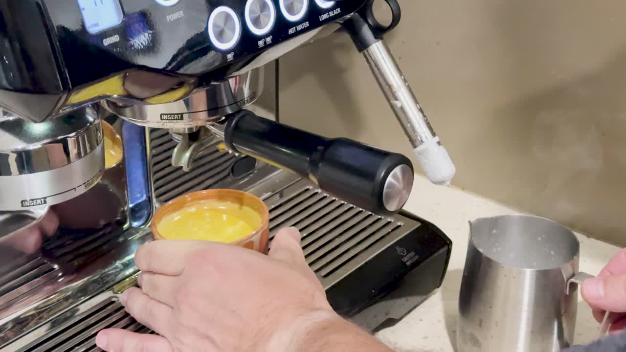Hands prepare coffee using a machine, steaming milk, and pouring into a cup. Bright kitchen lighting enhances the process
