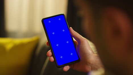 cerca sobre el hombro de un hombre que pasa la noche en casa sentado en el sofá mirando la pantalla azul del teléfono móvil