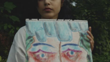 a woman with dark hair holds a drawing of a face