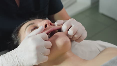 close up of recipient lying relaxed on spa bed eyes closed mouth open gently as professional in gloves dips fingertips into mouth massaging lips and jawline to enhance facial relaxation
