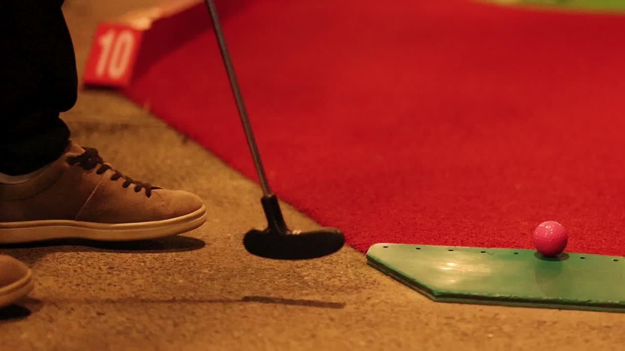 A close-up shot focusing on a person's feet and putter hitting a pink golf ball