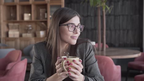 bella joven con gafas está sentada en un café, sosteniendo una taza de café y mirando la ventana