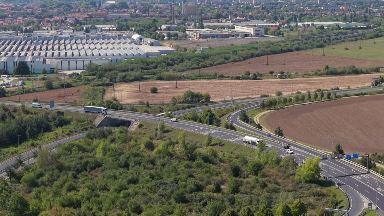 An aerial view shows a modern highway interchange surrounded by green fields and farmland. Trucks and cars travel along the road, a large industrial complex and urban area visible in the background