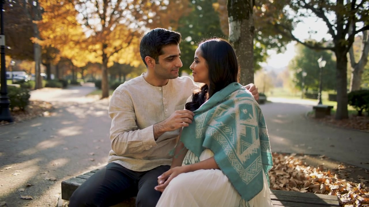 Lifting man in beige kurta draping teal shawl, warming woman in white dress on park bench