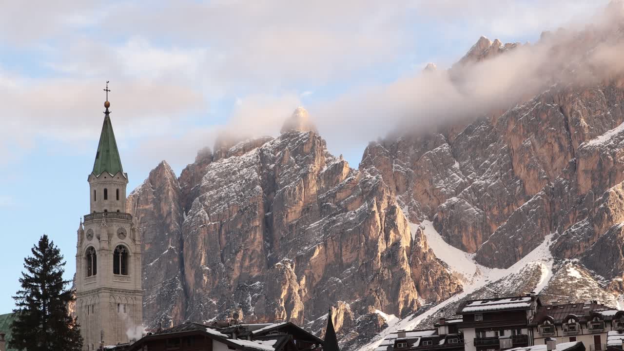 Cinematic shot of the iconic church bell tower of Cortina d'Ampezzo framed by the towering snowy peaks of the Dolomites. Alpine landmark in Italy