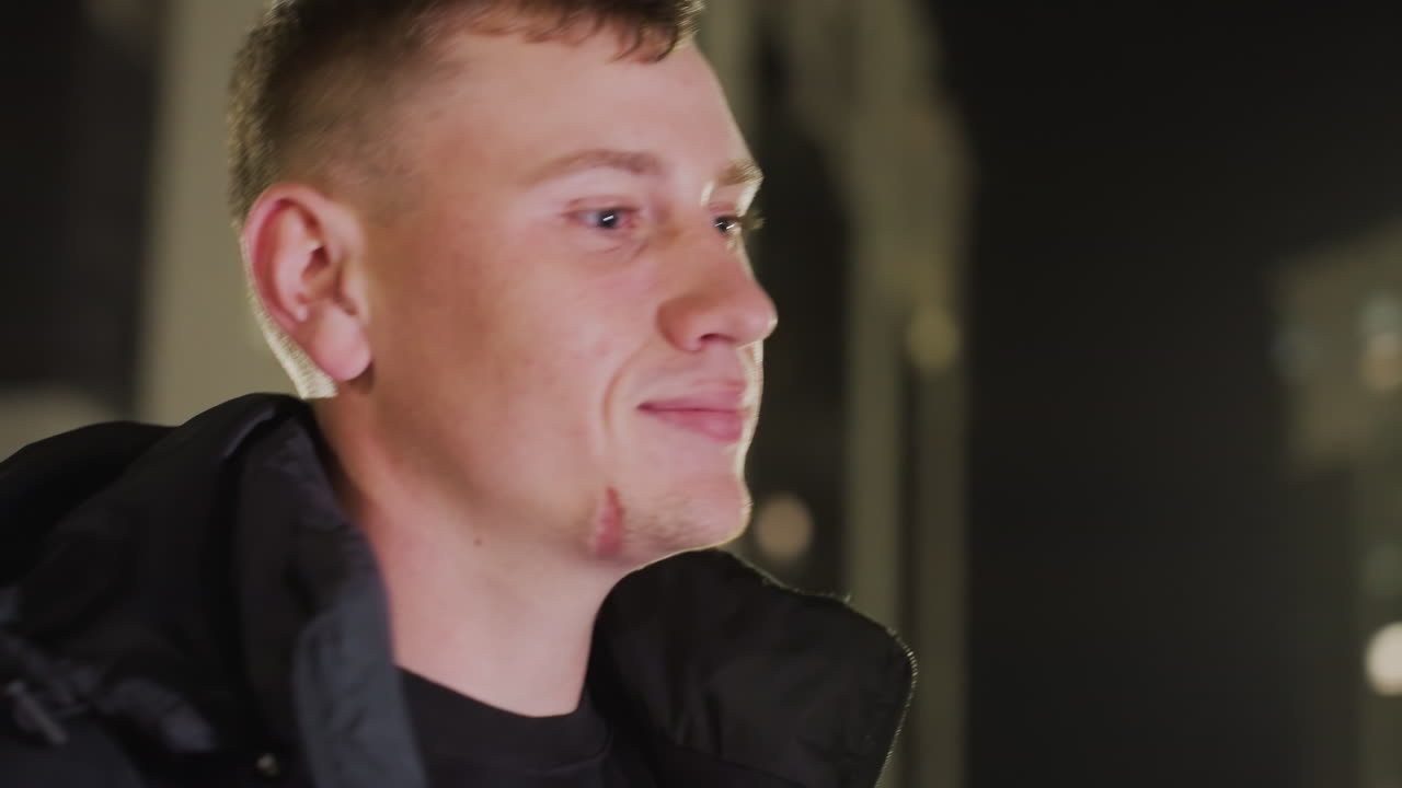 Young man wearing summer jacket walks and smiles warmly as he gently bends forward under soft glowing city lights in outdoor nighttime setting surrounded by blurred buildings