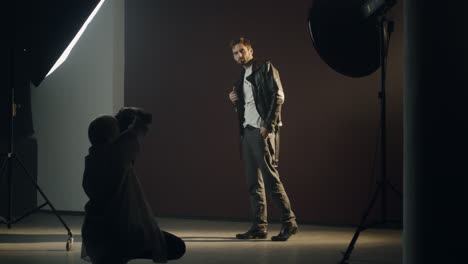 caucasian stylish young man posing to the camera of the female photographer in a dark studio during photosession