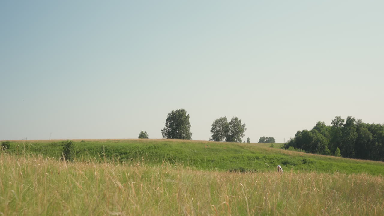 Wide distant view of person wearing white hat walking across grassy meadow toward low hill under clear blue sky with scattered trees and peaceful green landscape