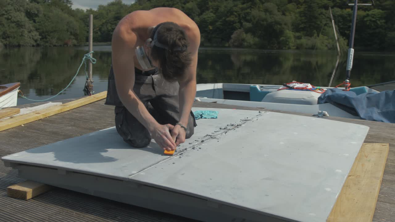 Young shirtless man scraping off excess mastic sealant on plywood engine cover, Medium Wide Shot