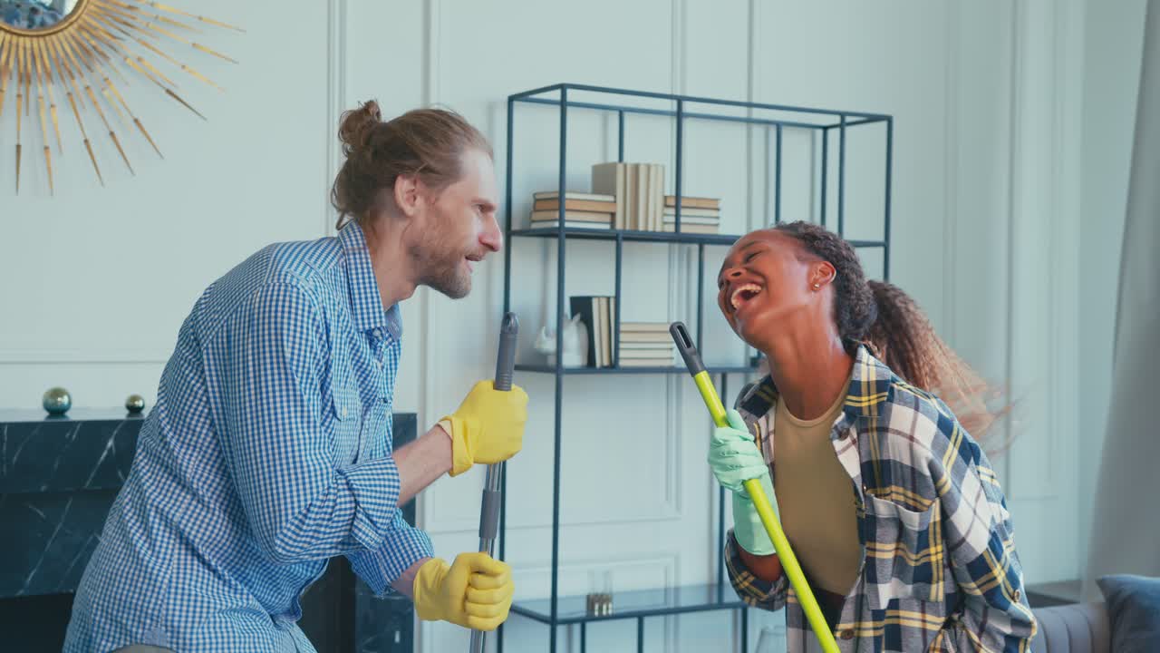 una pareja alegre comparte un momento lúdico mientras limpian su sala de estar juntos.