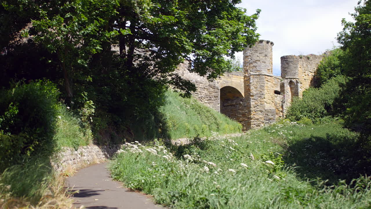 A medieval castle gate entrance in Scarborough, North Yorkshire, England, framed by tall trees and summer light. A majestic pathway bordered by stone walls and greenery