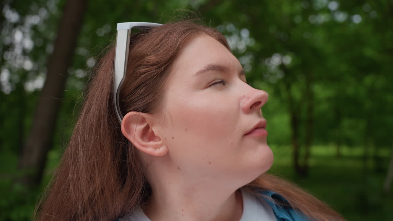 Close up of lady in casual outfit sitting outdoors on wooden bench sipping water from clear plastic bottle, relaxing under green trees on warm summer day, enjoying refreshing drink
