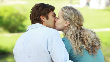 a man and woman sit in the park embraced as they kiss each other