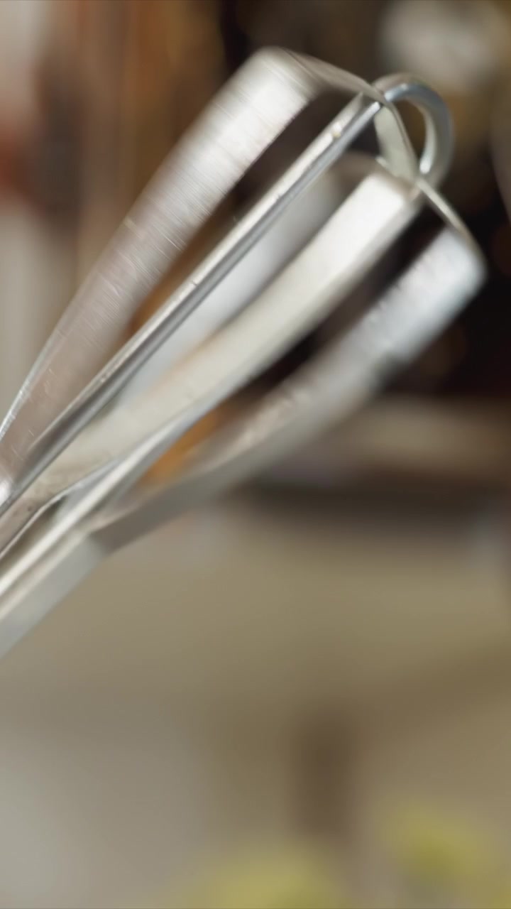 Close up of metal whisk rotating slowly in focus emphasizing. Continuous motion highlights metallic construction details. Background suggests kitchen environment with warm tones