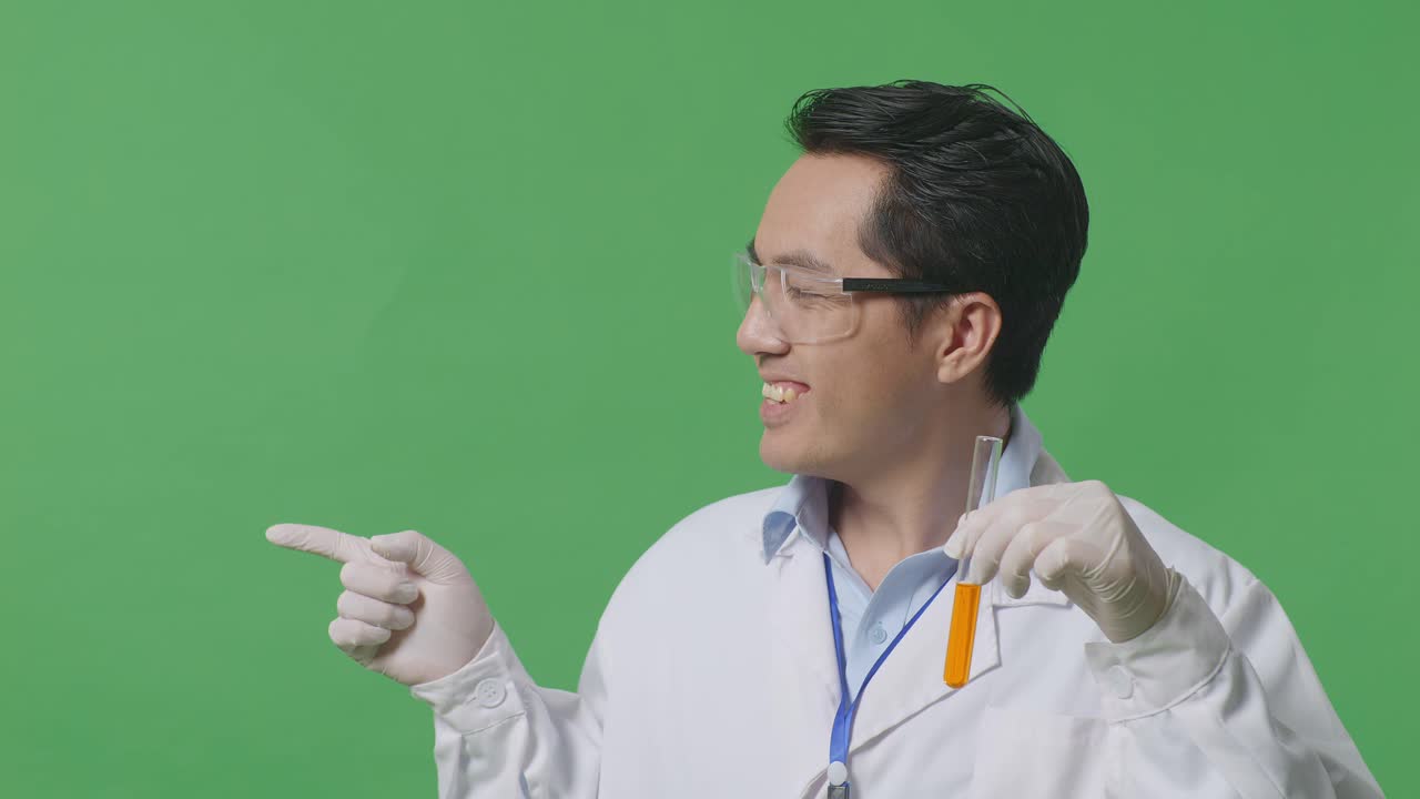 Close Up Of Asian Man Scientist With Orange Liquid In The Test Tube Smiling And Pointing To Side While Standing On The Green Screen Background In The Laboratory