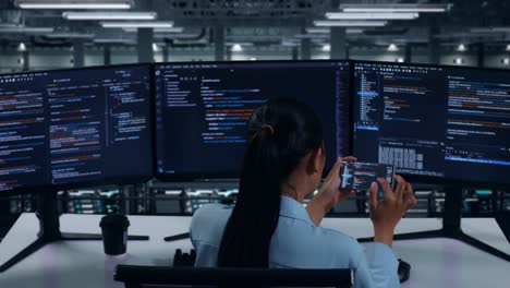 vista trasera de una desarrolladora asiática trabajando con un teléfono móvil mientras escribe código con múltiples pantallas de computadora en un centro de datos