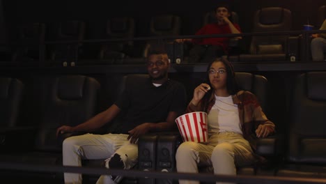 mixed raced young couple sitting in movie theatre watching film and taking popcorn