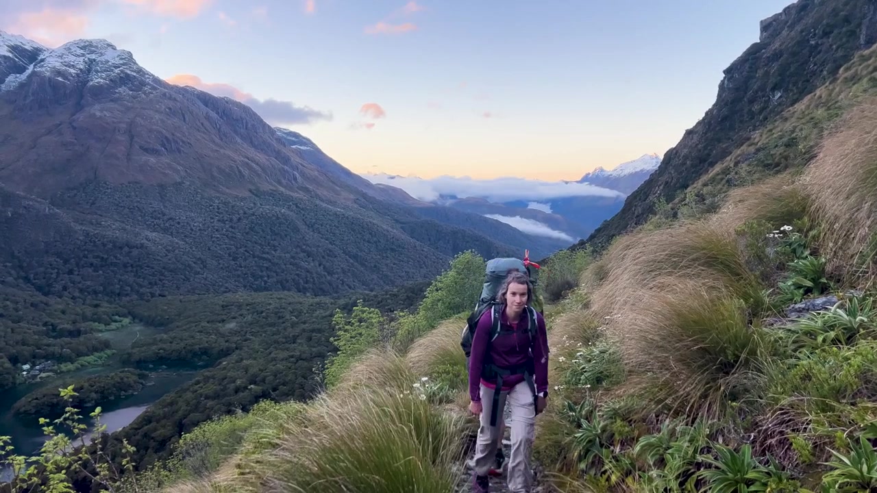 A tramper with a full pack climbs the Routeburn Track in New Zealand at dawn, tussock crowding the stony path and cloud filling the valley far below. Static wide shot
