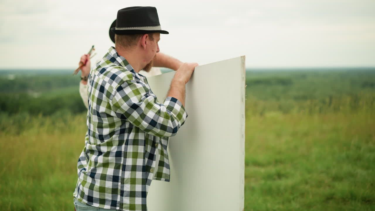 un artista con un sombrero negro y una camisa a cuadros sostiene una gran tabla blanca al aire libre en un campo cubierto de hierba. una mujer con un vestido blanco, con un pincel de pintura, está cerca observando la escena con interés