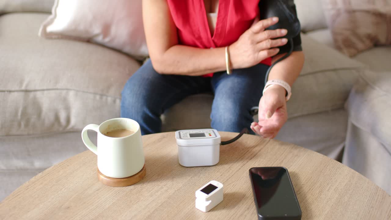 Donna anziana asiatica in camicetta rossa che avvolge il bracciale, collega il monitor e controlla la lettura della pressione sanguigna con un segnale acustico.