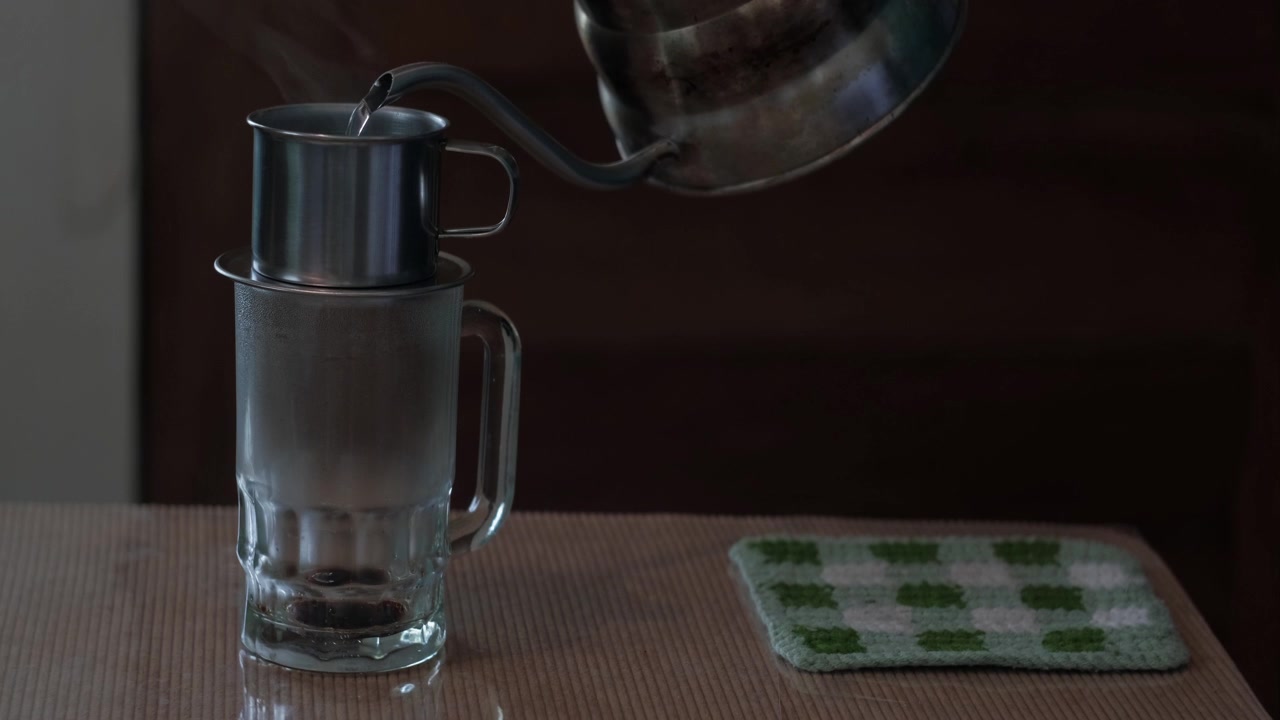 A gooseneck kettle pouring hot water into a Vietnamese phin filter over a glass mug to brew coffee indoors - medium static shot