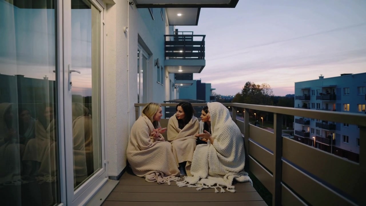 Tre donne rannicchiate per il calore, che si avvicinano e parlano su un balcone mentre il crepuscolo si intensifica, con coperte.